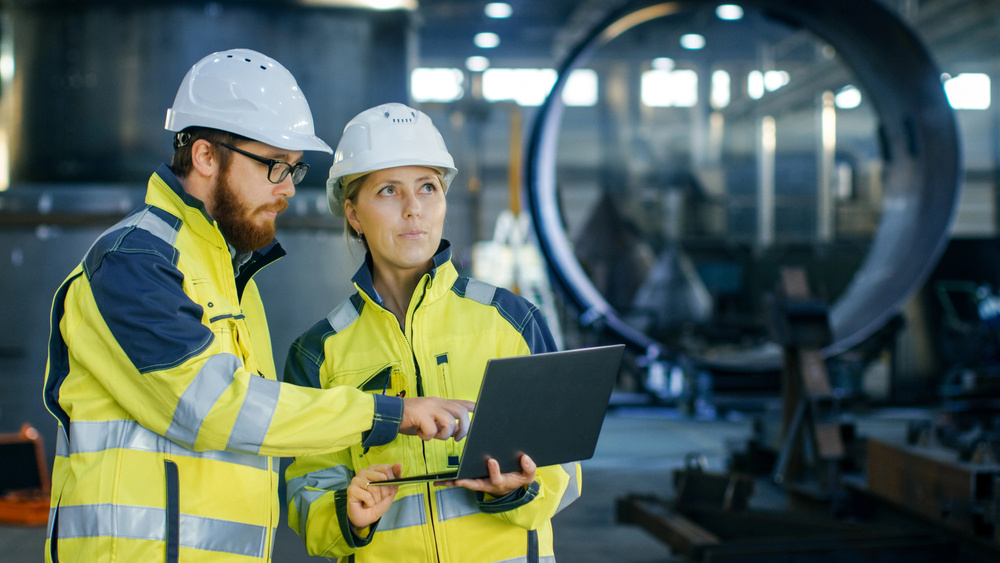 Two workers wearing safety helmets and vests, analyzing data on a computer in an industrial setting.